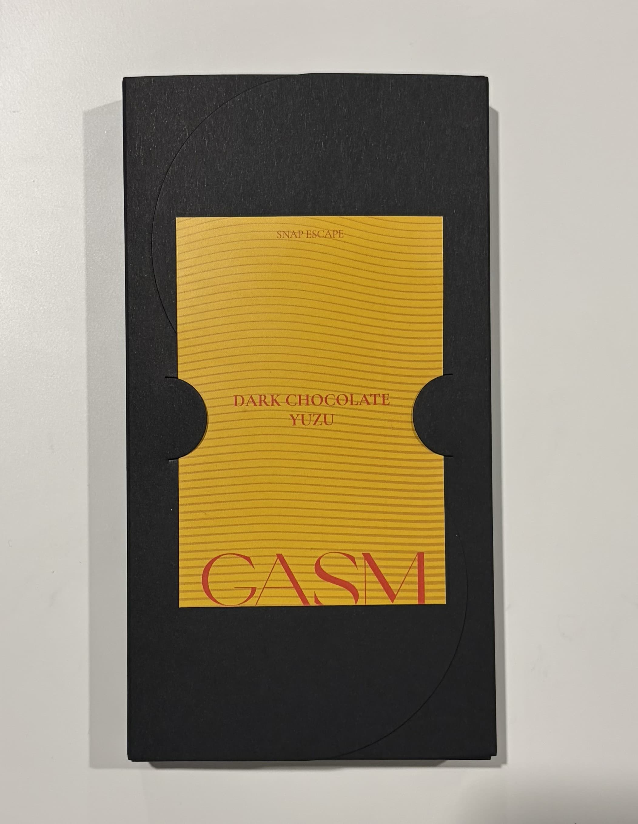 A GASM dark chocolate yuzu bar in its black sleeve, lying flat on a
                    pale worktop under ordinary room light.