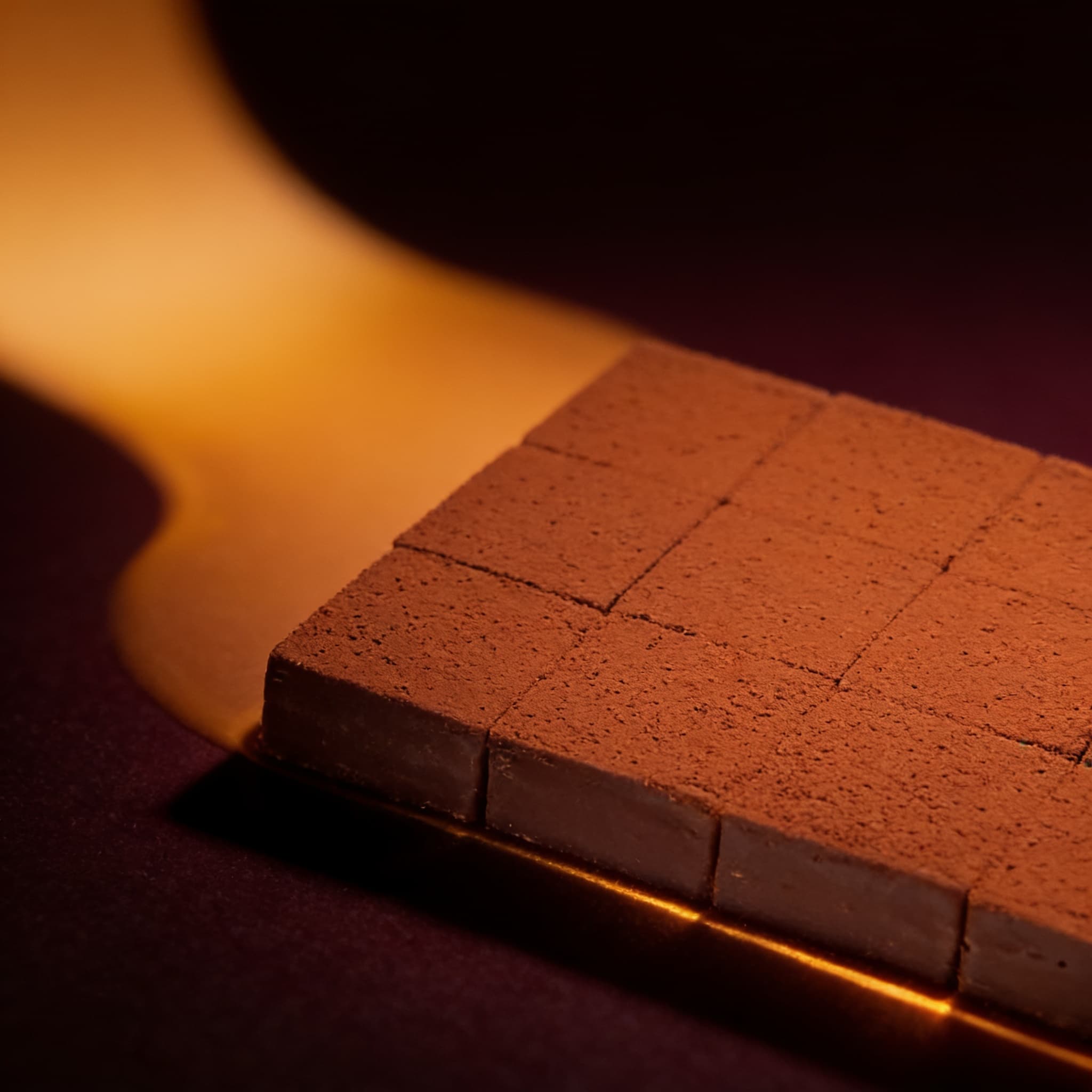 A cocoa-dusted chocolate slab scored into squares, raked by a warm gold light against a deep plum ground.