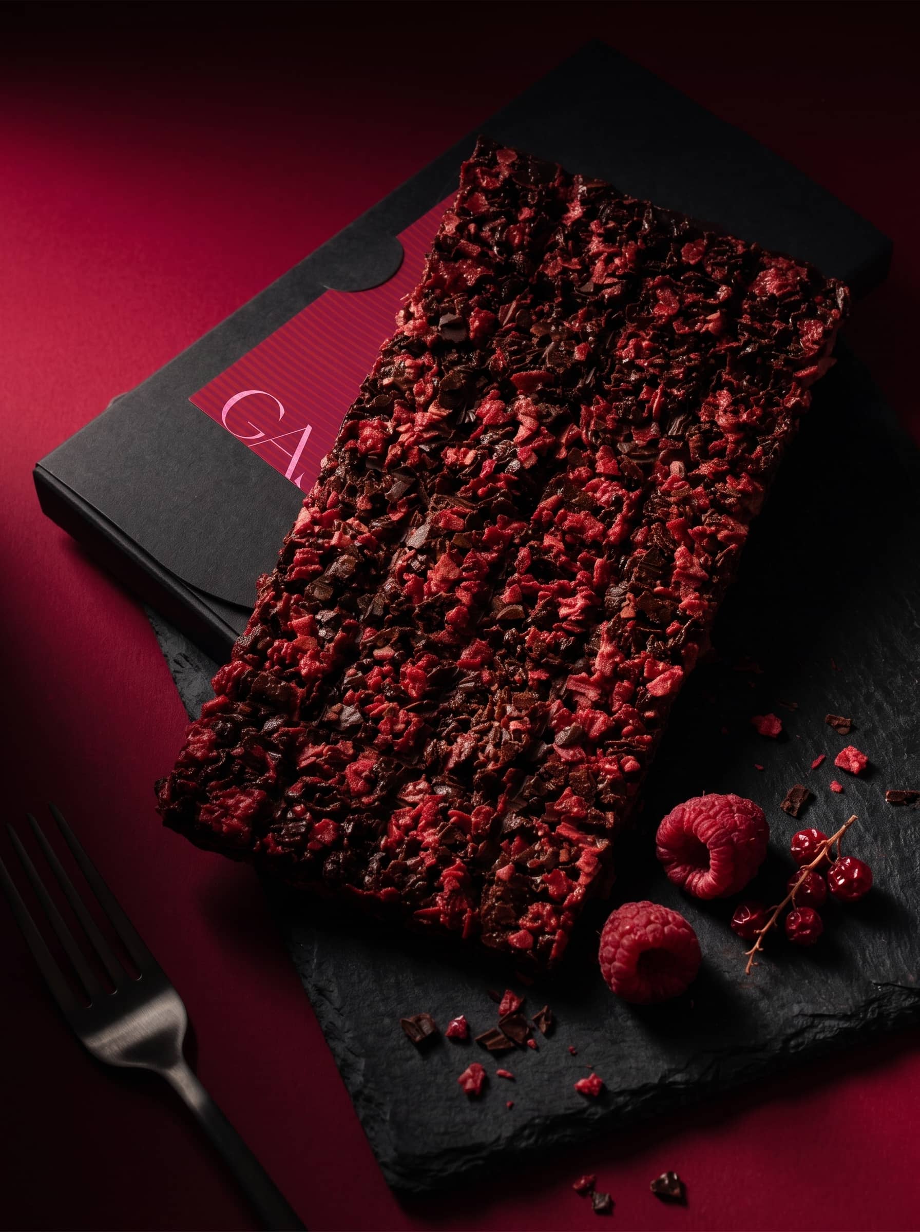 A raspberry-topped chocolate slab on slate beside its black GASM box, with fresh raspberries, redcurrants and a fork.